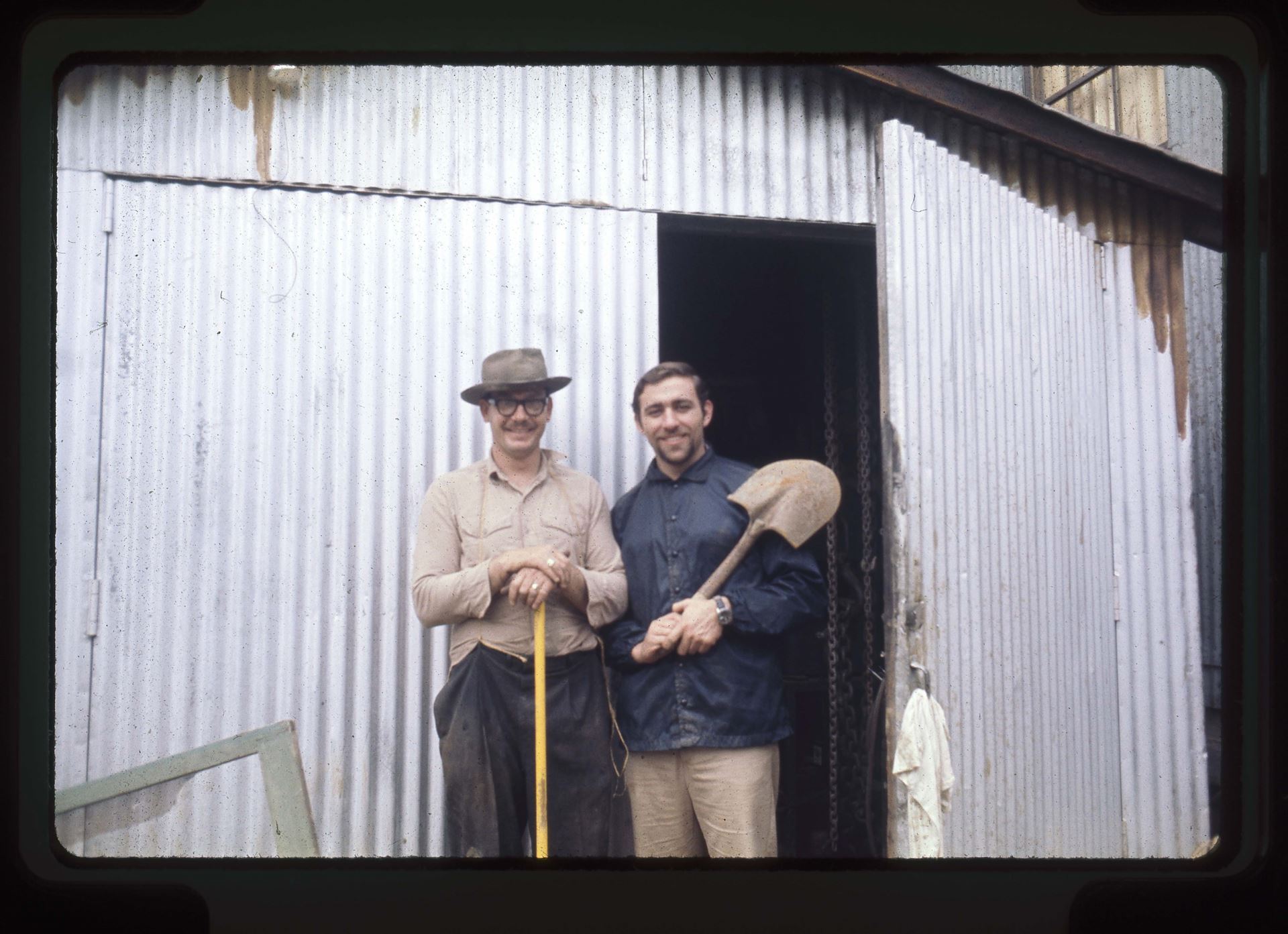 Michael Rubel and Dwayne Hunn with farm tools, ca. 1969-01 35mm slides (photographs), hunn-c2019-2-11-02~006, Michael Rubel and Dwayne Hunn with shovels during flood mud management, standing in front of Tractor Shed, "JAN 69" stamped on slide mount, Rubelia, Rubel Castle, Rubel Pharms, Rubel Farms, ca. January, 1969.